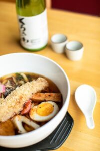 A bowl of tempura udon noodle soup and a bottle of sake with two cups at Shema Sushi Japanese restaurant in Rochester NY