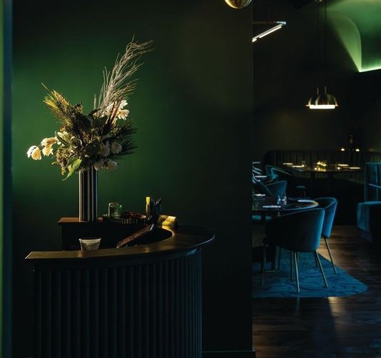 The host stand at Patron Saint with a luxurious simply chandelier hanging and flowers in a slender vase all in low yellow lighting with dark emerald green paint on the walls in Rochester, N.Y.