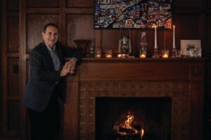 George Conboy, chairman of Brighton Securities, poses and leans on wooden fireplace mantel with several glass jars with lit candles and a mosaic artwork hung in center of the fireplace in Rochester, N.Y.