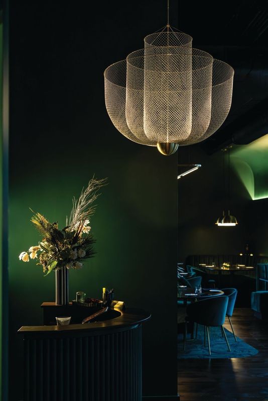 The host stand at Patron Saint with a luxurious simply chandelier hanging and flowers in a slender vase all in low yellow lighting with dark emerald green paint on the walls in Rochester, N.Y. 