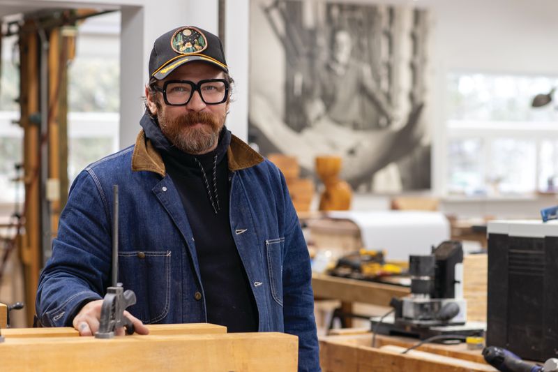 Artist Christian Wargo posing in a woodshop wearing a hat, black hoodie, and jean jacket in Rochester, N.Y.