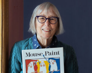 Author Ellen Stoll Walsh smiling and holding up her children's book Mouse Paint.